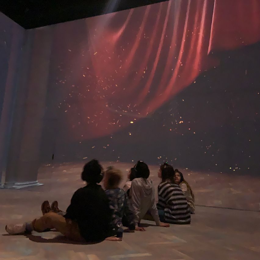 Eine Gruppe von Menschen sitzt auf dem Boden in einem Raum mit einem roten Vorhang. Sie sehen sich eine Show mit einem funkelnden Effekt an.