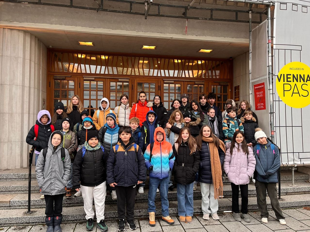 Eine Gruppe junger Menschen in Winterkleidung posiert für ein Foto vor einem Gebäude mit Glastüren.