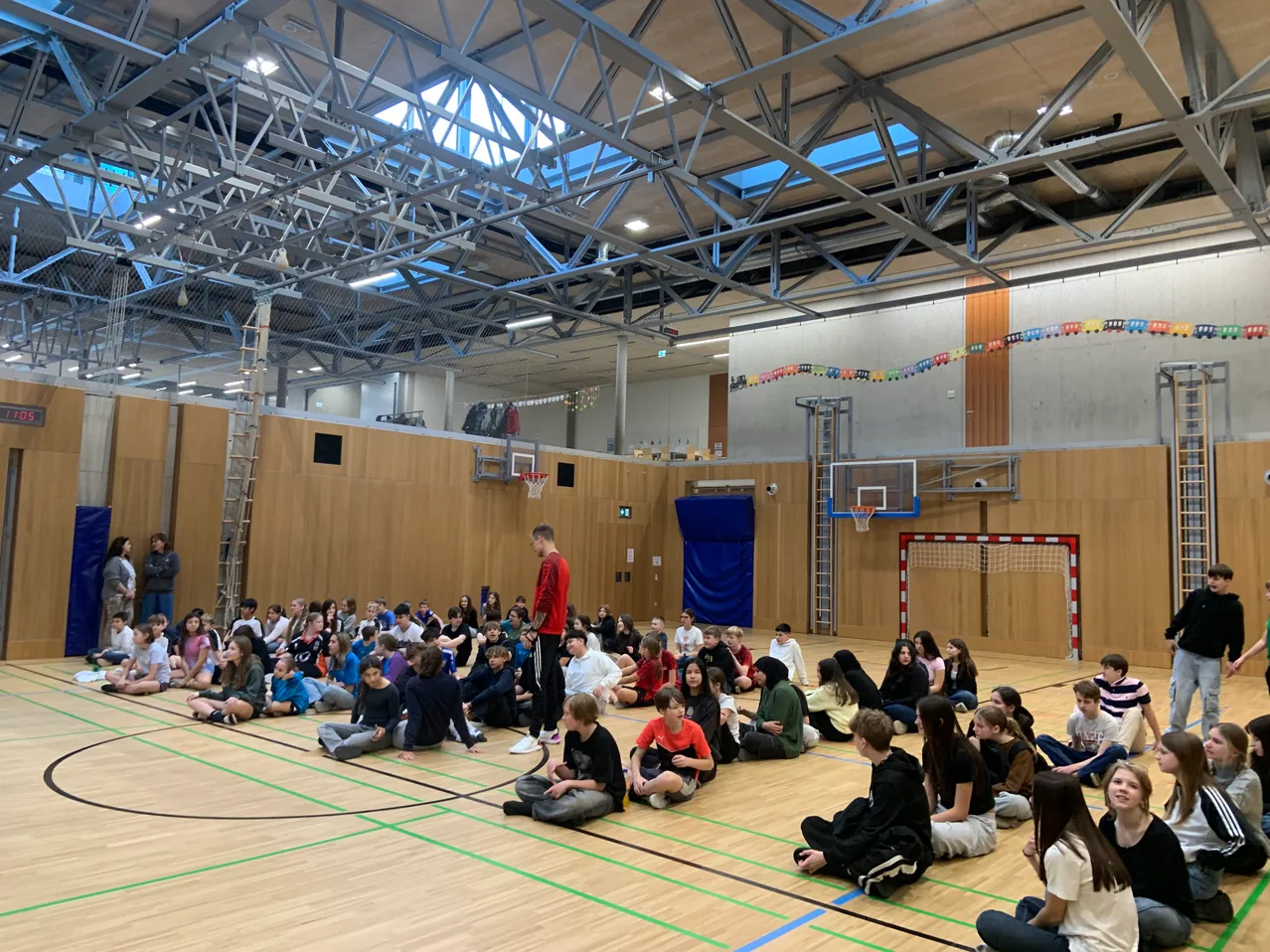 Eine Gruppe von Menschen sitzt auf dem Boden einer Turnhalle und hört einem Mann in einem roten Shirt zu, der vor ihnen steht. An den Wänden befinden sich Basketballkörbe und eine Leiter lehnt an einer der Wände.