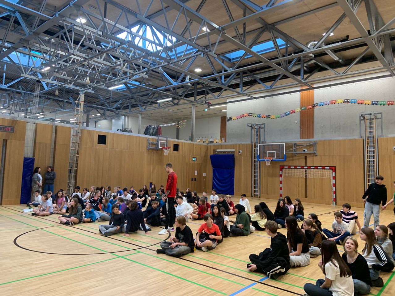 Eine Gruppe von Menschen sitzt auf dem Boden einer Turnhalle und hört einem Mann in einem roten Shirt zu, der vor ihnen steht. An den Wänden befinden sich Basketballkörbe und eine Leiter lehnt an einer der Wände.