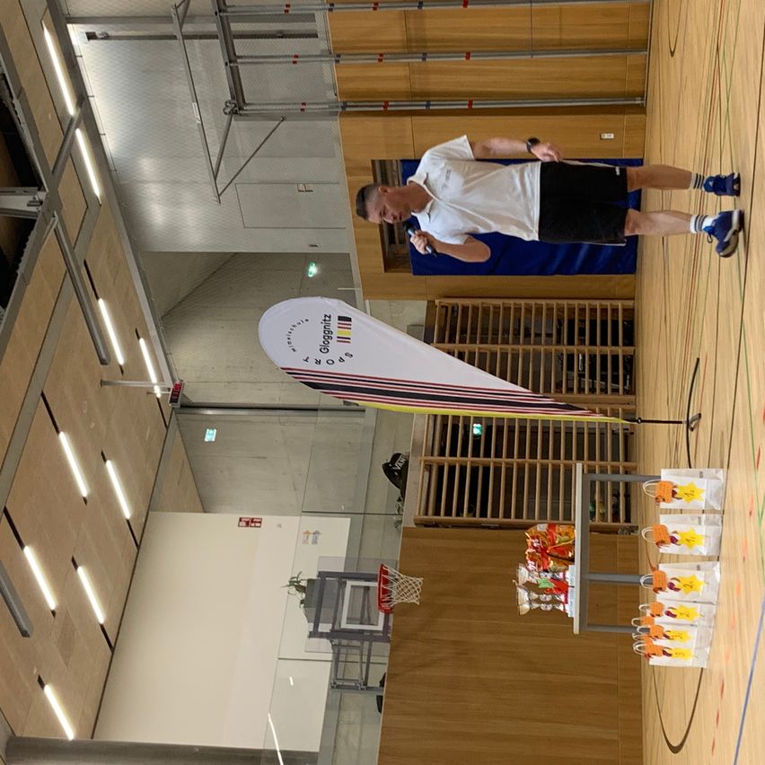 Ein Mann spricht in ein Mikrofon und steht in einer Turnhalle. Ein weißer Banner mit bunten Streifen und dem Namen 'Gognitz' ist in der Nähe. Ein Basketballkorb ist sichtbar, und verschiedene Gegenstände stehen auf einem Tisch.