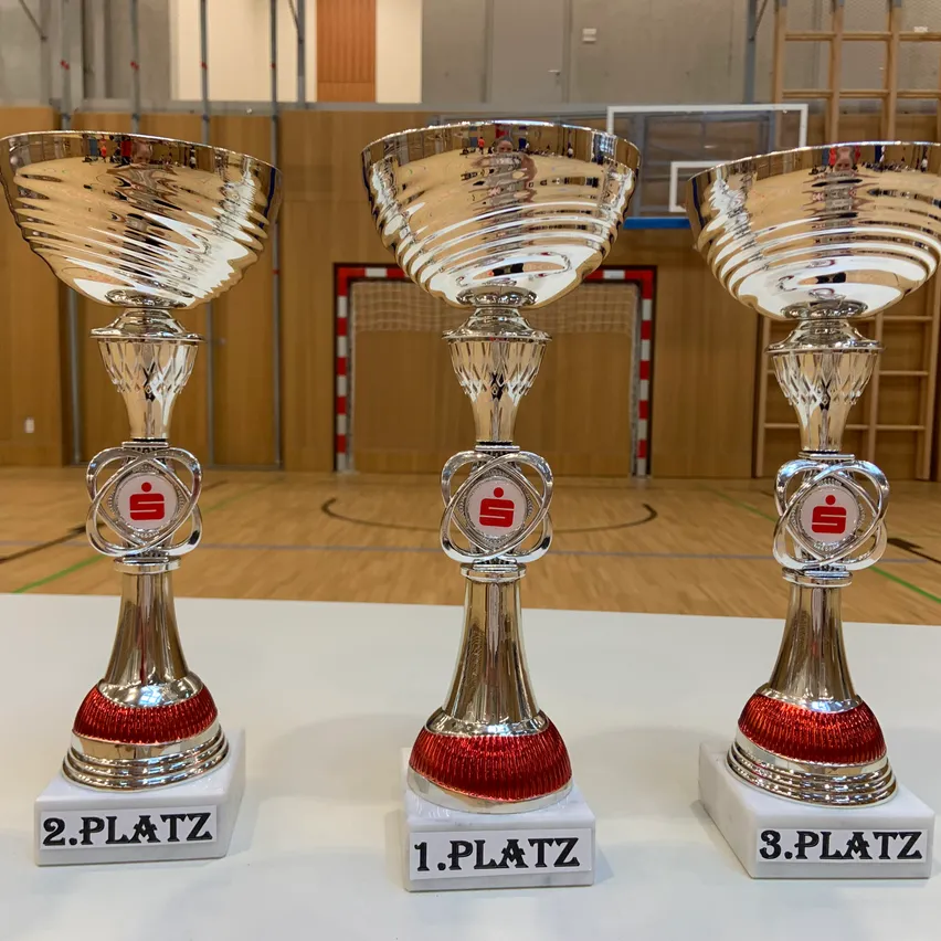 Drei silberne Pokale mit roten Akzenten stehen auf einer weißen Oberfläche. Jeder Pokal hat eine andere Positionsbezeichnung: '2. Platz', '1. Platz' und '3. Platz'. Sie befinden sich in einer Sporthalle mit Holzboden und einem Basketballplatz im Hintergrund.
