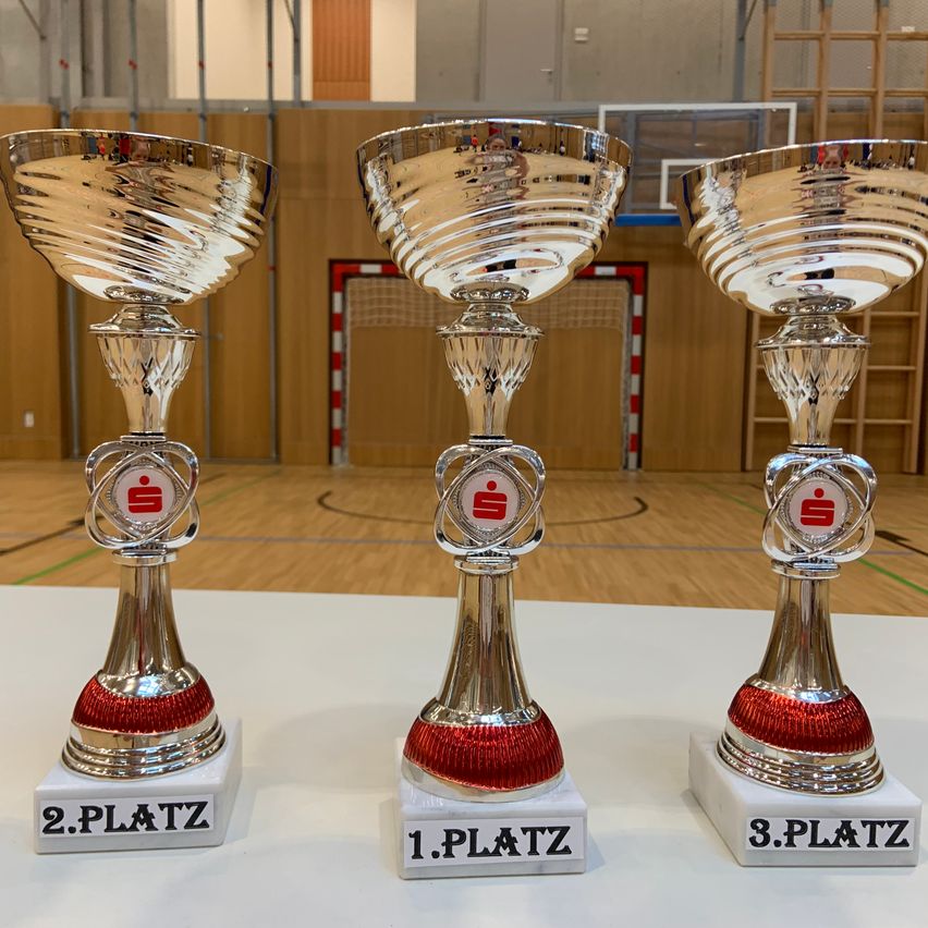 Drei silberne Pokale mit roten Akzenten stehen auf einer weißen Oberfläche. Jeder Pokal hat eine andere Positionsbezeichnung: '2. Platz', '1. Platz' und '3. Platz'. Sie befinden sich in einer Sporthalle mit Holzboden und einem Basketballplatz im Hintergrund.