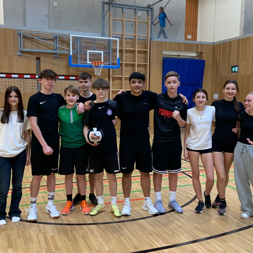 Eine Gruppe junger Menschen steht lächelnd auf einem Basketballplatz. Ein Junge hält einen Fußball. Im Hintergrund ist ein Basketballkorb zu sehen.