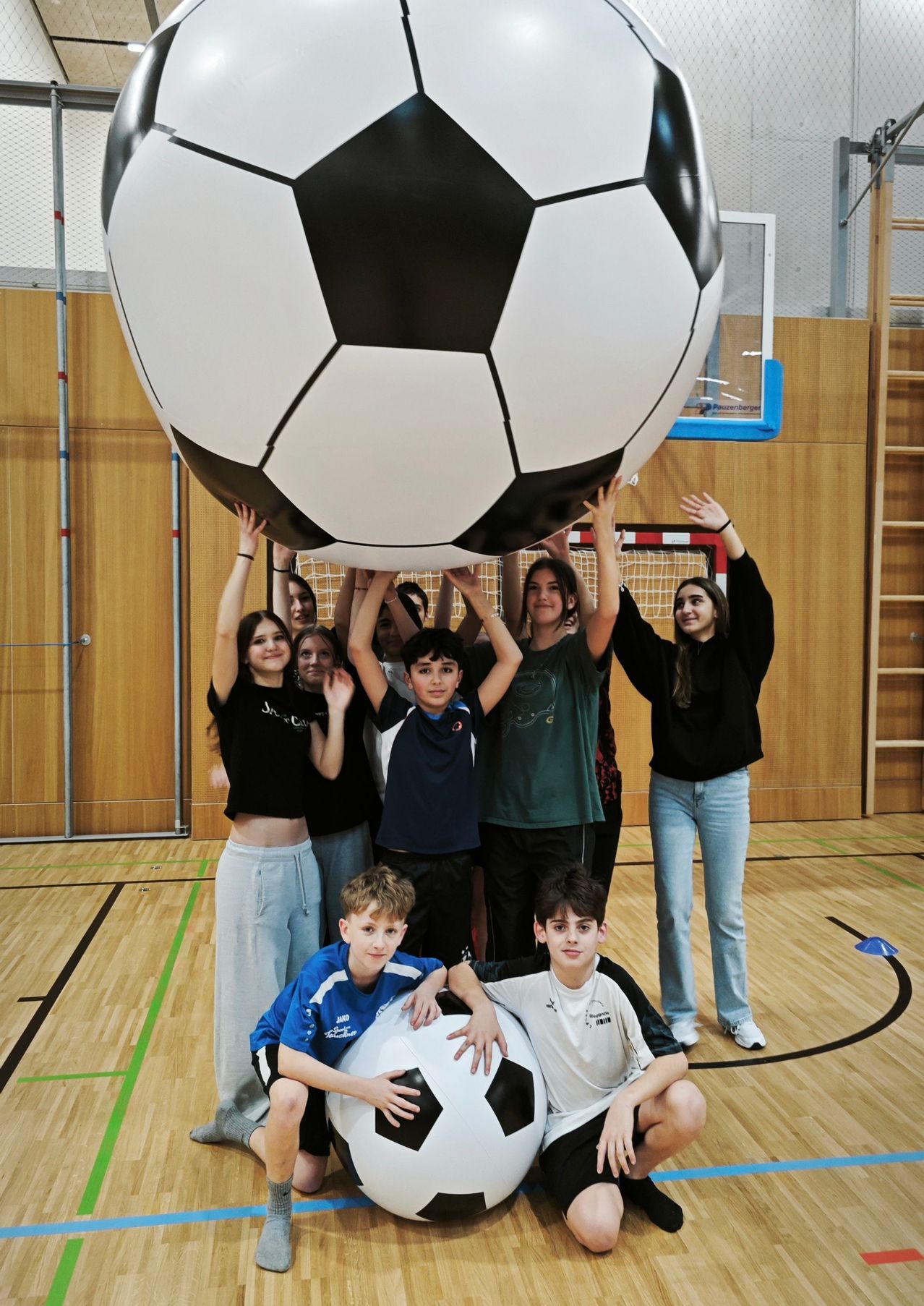Eine Gruppe junger Menschen hält einen riesigen aufgeblasenen Fußball in einer Turnhalle hoch. Sie lächeln und posieren für ein Foto.