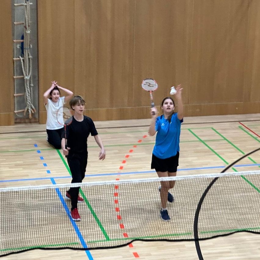 Drei Personen spielen Badminton auf einem Hallenspielfeld. Zwei sind aktiv beteiligt, während die dritte hinter ihnen hockt. Das Spielfeld hat einen Holzboden und mehrfarbige Markierungen.
