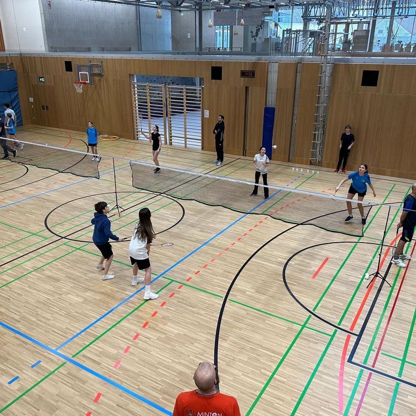 Eine Gruppe von Menschen spielt Badminton auf einer Indoor-Spielfläche. Die Spielfläche ist hölzern mit bunten Markierungen. Einige Spieler schlagen den Federball, während andere zuschauen.