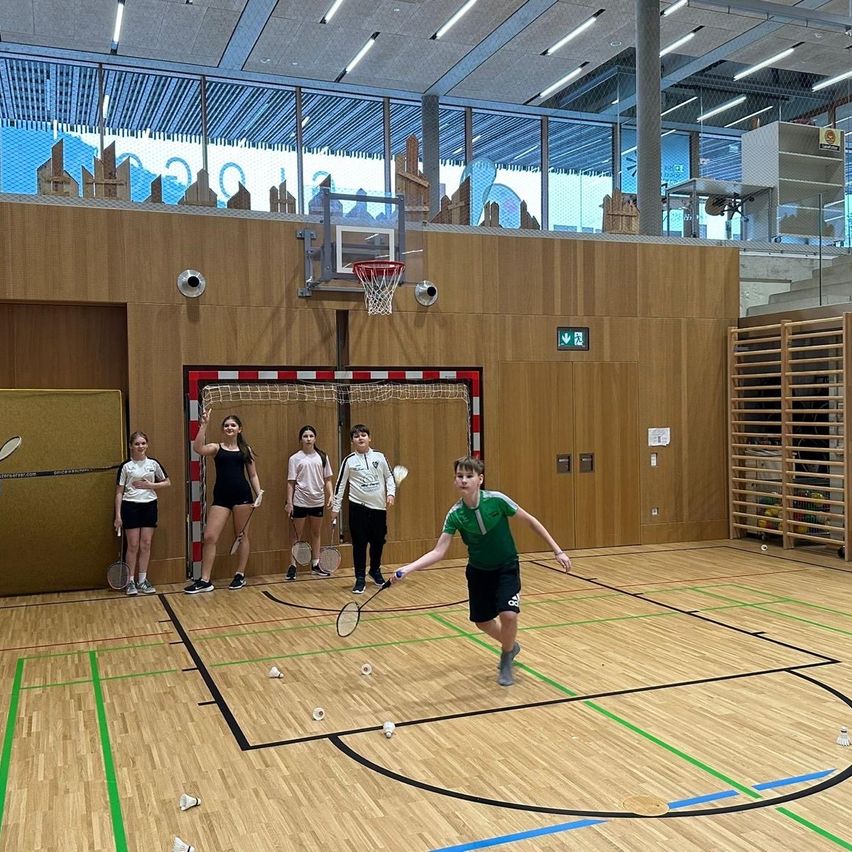 Eine Gruppe junger Menschen spielt Hallenbadminton, ein Junge ist dabei, den Federball zu schlagen, während andere hinter einem Netz zusehen.