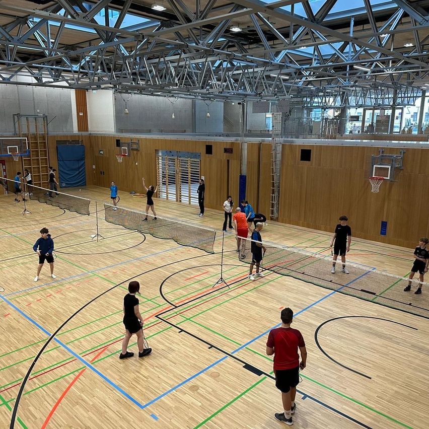 Innenhalle mit mehreren Badmintonnetzen. Mehrere Personen spielen in Teams Badminton, einige schauen zu. Holzwände und eine Metalldecke.
