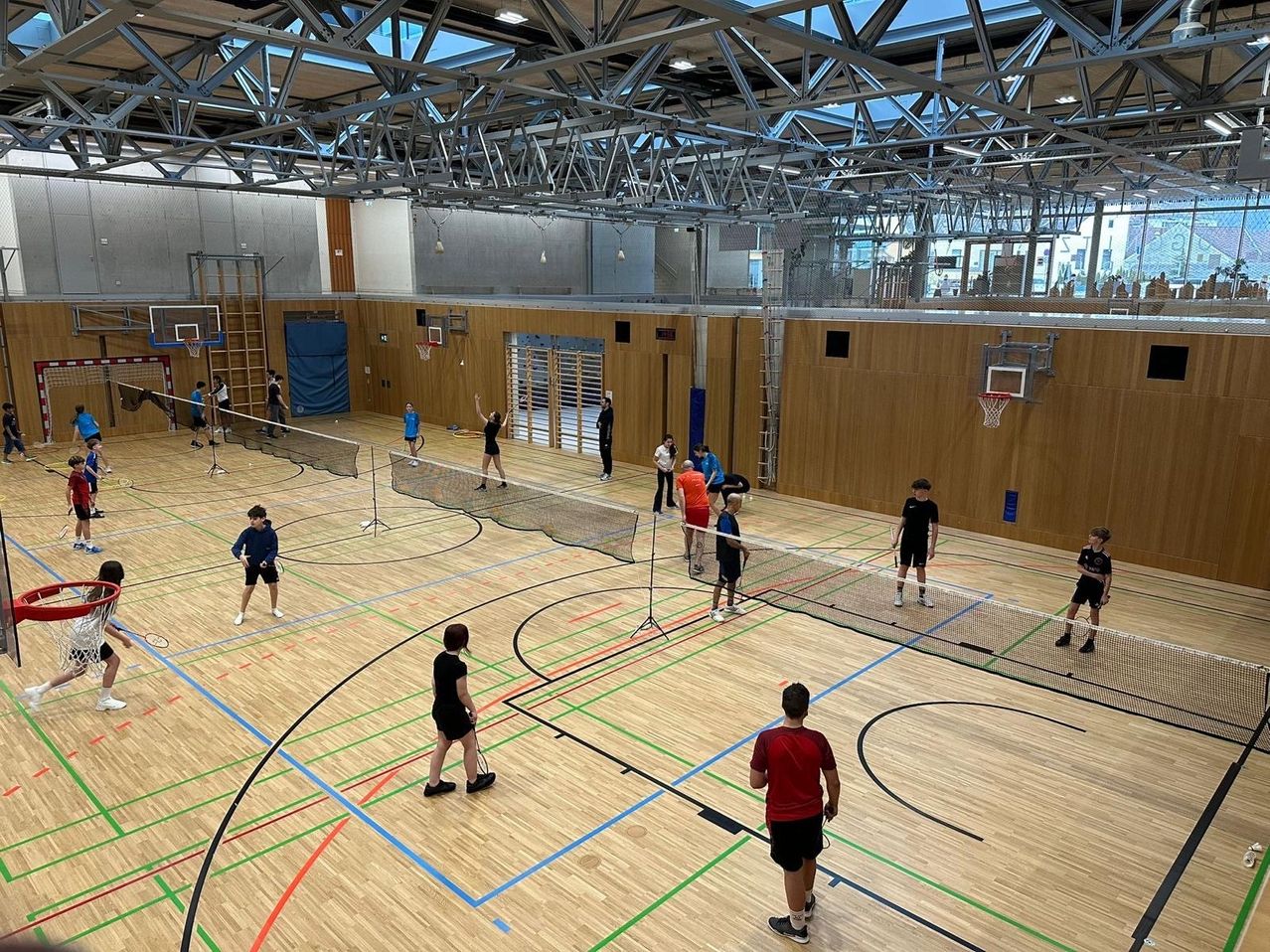 Innenhalle mit mehreren Badmintonnetzen. Mehrere Personen spielen in Teams Badminton, einige schauen zu. Holzwände und eine Metalldecke.