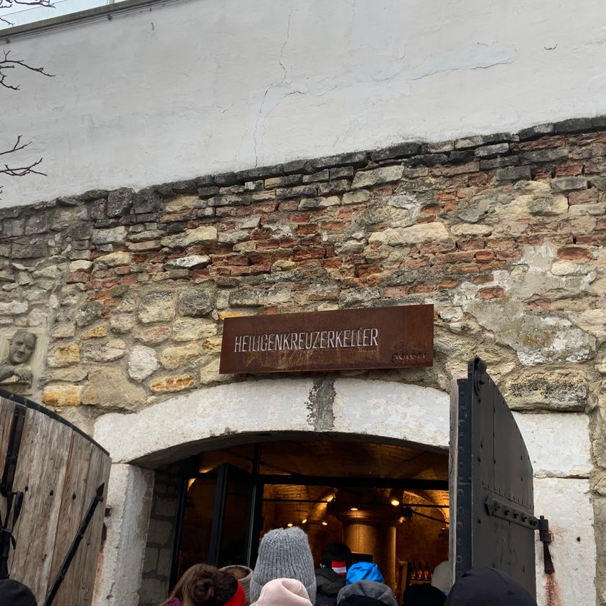 Ein Steingebäude mit einer Holztür und einem Schild mit der Aufschrift Heiligenkreuzerkeller. Vor dem Eingang stehen einige Menschen.