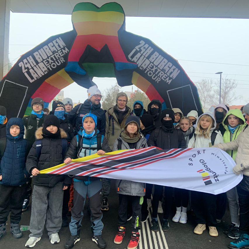 Eine Gruppe von Menschen in Winterkleidung steht unter einem farbenfrohen Bogen mit der Aufschrift '24h Burgen Land Extrem Tour' und hält ein Banner mit den Worten 'Sport Gloggnitz'. Sie sind draußen, wobei einige Bäume und eine Straßenlaterne im Hintergrund sichtbar sind.
