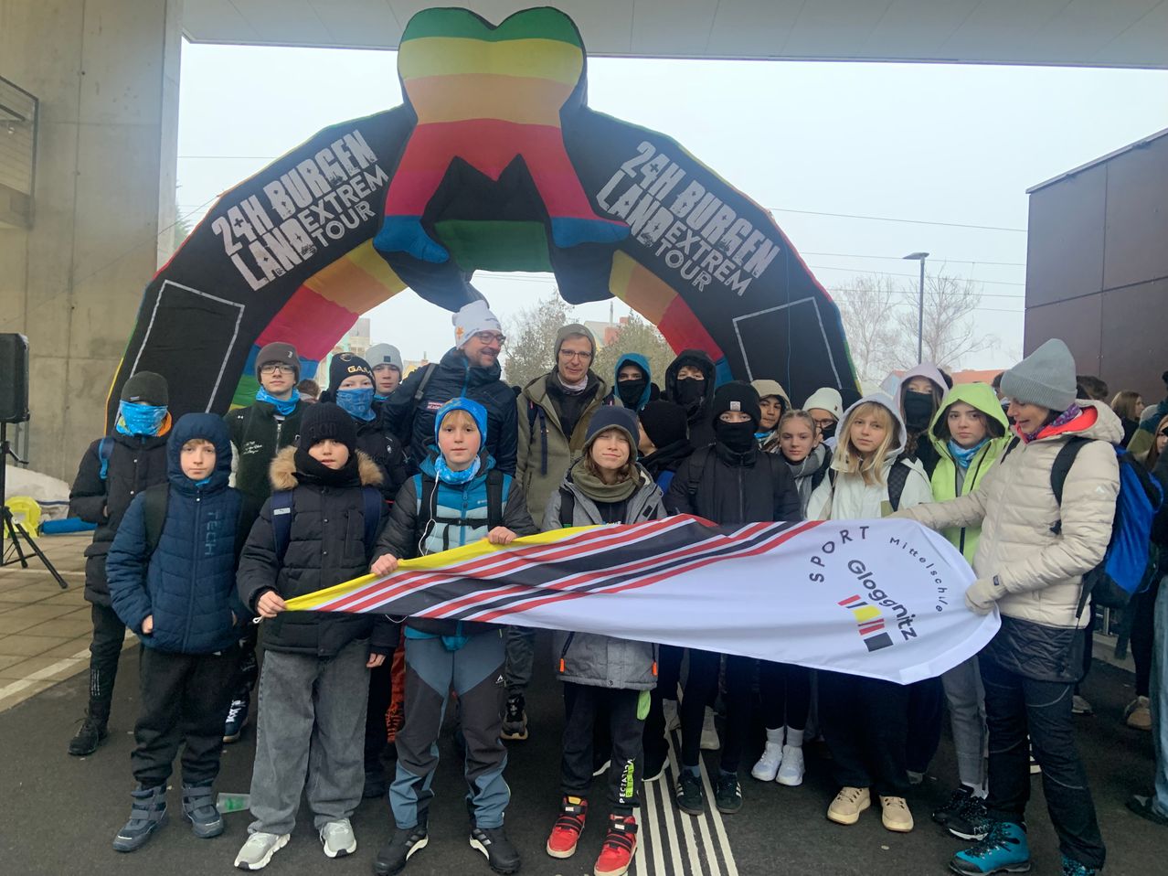 Eine Gruppe von Menschen in Winterkleidung steht unter einem farbenfrohen Bogen mit der Aufschrift '24h Burgen Land Extrem Tour' und hält ein Banner mit den Worten 'Sport Gloggnitz'. Sie sind draußen, wobei einige Bäume und eine Straßenlaterne im Hintergrund sichtbar sind.