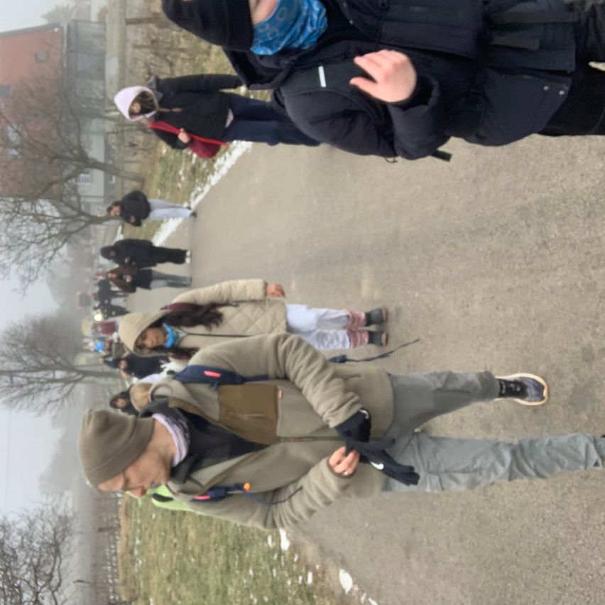 Eine Gruppe von Menschen läuft auf einem nebligen Pfad. Sie sind in Winterkleidung gekleidet. Eine Person trägt einen Rucksack und eine andere einen Hut. Der Boden ist mit Schnee bedeckt. An den Seiten stehen Bäume.