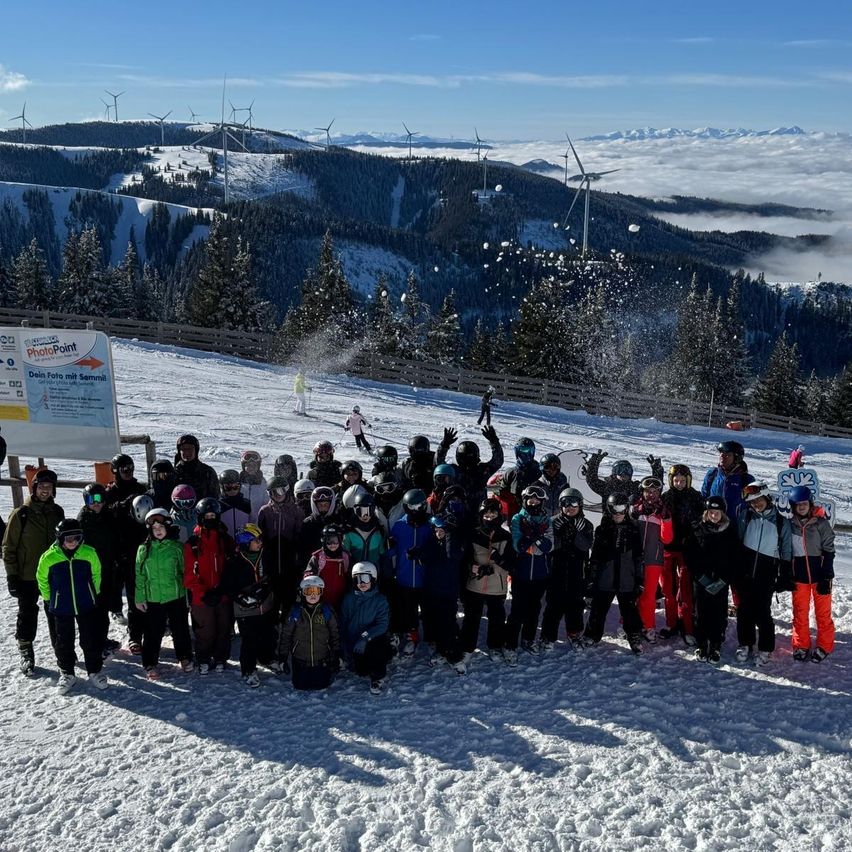 Eine Gruppe von Menschen posiert für ein Foto auf einem verschneiten Hang, mit Windturbinen und Bergen im Hintergrund.