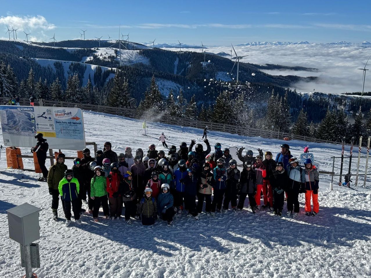 Eine Gruppe von Menschen posiert für ein Foto auf einem verschneiten Hang, mit Windturbinen und Bergen im Hintergrund.