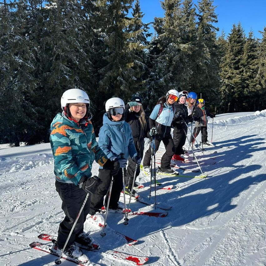 Eine Gruppe von Skifahrern in Winterkleidung steht in einer Reihe auf einem verschneiten Hang mit Bäumen im Hintergrund.