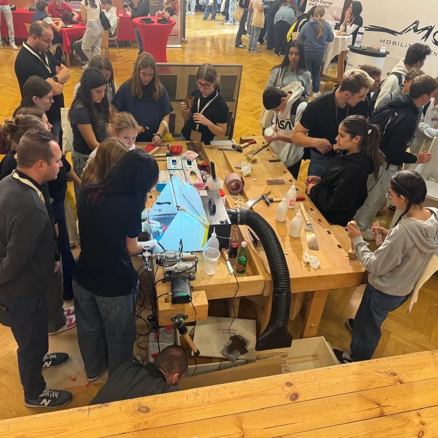 In einem Workshop versammeln sich mehrere Personen um einen Holztisch und arbeiten an verschiedenen Objekten. Der Tisch ist mit Werkzeugen und Materialien gefüllt. Einige Personen stehen, andere sitzen. Der Raum hat Holzböden und -wände, mit Tischen und Stühlen im Hintergrund.