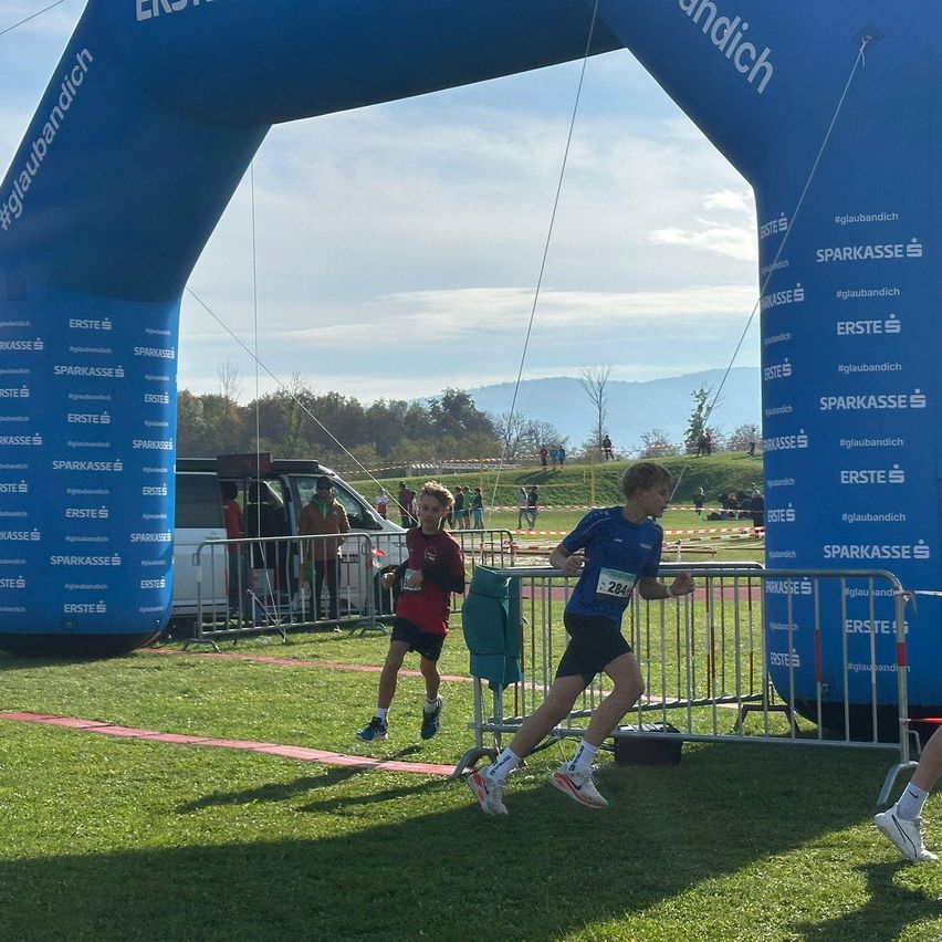 Zwei Kinder laufen bei einem Marathon und passieren einen blauen Bogen mit der Aufschrift 'ERSTE SPARKASSE'. Dahinter ist ein Van geparkt und eine Person steht am Zaun. Bäume und Berge sind im Hintergrund.
