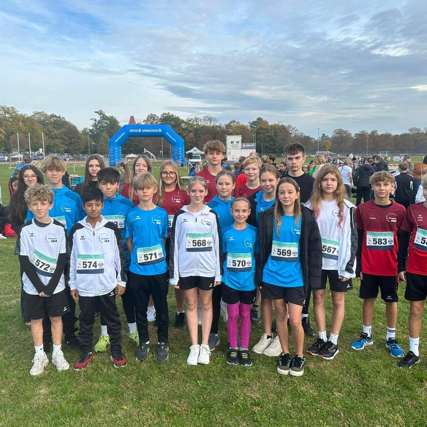 Eine Gruppe von Kindern und Jugendlichen in Sportkleidung steht auf einem Rasenfeld, wahrscheinlich nach einem Rennen. Sie tragen Startnummern und haben verschiedene Ausdrücke. Hinter ihnen befindet sich ein blauer Bogen und eine Menschenmenge.