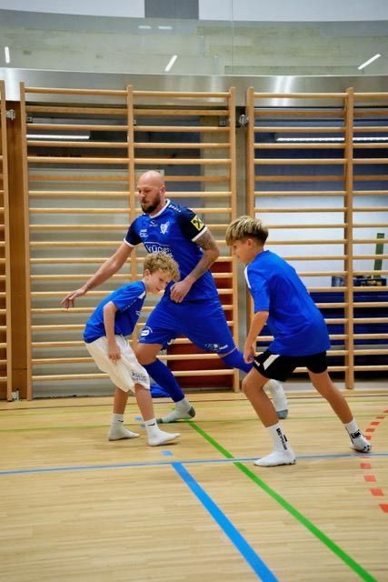 A man and two boys in blue uniforms are playing soccer on a wooden floor with blue and green markings.