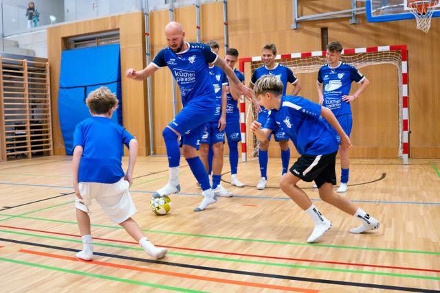 A group of soccer players, both adults and children, are playing on an indoor court. The players are wearing blue uniforms.