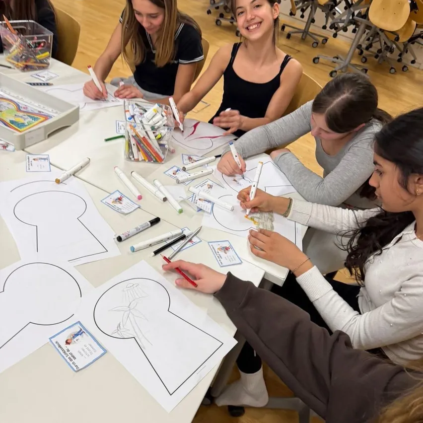 Eine Gruppe von Mädchen arbeitet an einem Projekt, bei dem sie Papierausschnitte auf einem Tisch ausmalen. Sie haben Marker und andere Malutensilien um sich herum verteilt.