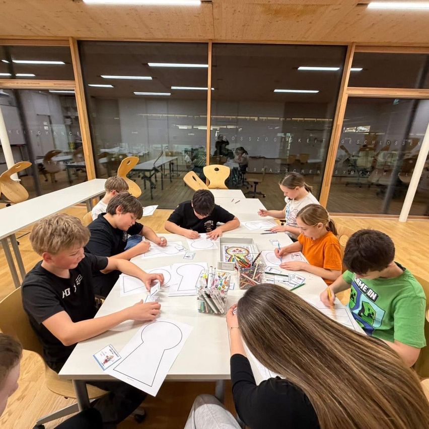Eine Gruppe von Kindern sitzt um einen Tisch in einem Klassenzimmer herum und färbt ein Schlüsselloch aus. Sie sind mit einer Malaktivität beschäftigt und es liegen viele Buntstifte auf dem Tisch. Das Klassenzimmer hat Glasfenster und der Raum ist gut beleuchtet.