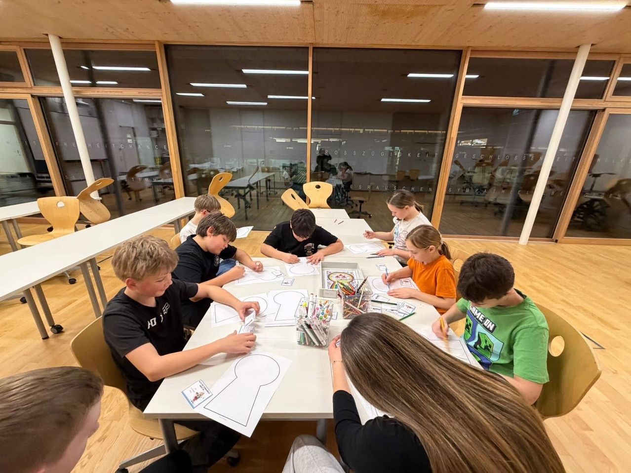 Eine Gruppe von Kindern sitzt um einen Tisch in einem Klassenzimmer herum und färbt ein Schlüsselloch aus. Sie sind mit einer Malaktivität beschäftigt und es liegen viele Buntstifte auf dem Tisch. Das Klassenzimmer hat Glasfenster und der Raum ist gut beleuchtet.
