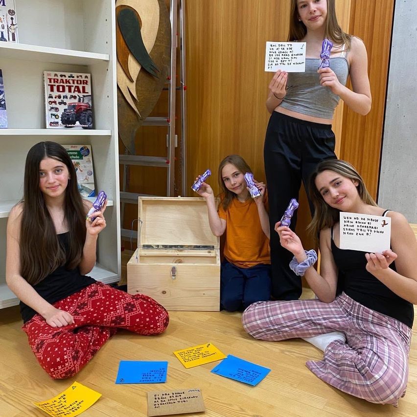 Vier junge Mädchen sitzen auf dem Boden in einem Zimmer, halten Schokoladen und Schilder mit Botschaften hoch. Eine hält eine Schachtel. Eine hölzerne Leiter und Bücher sind im Hintergrund.