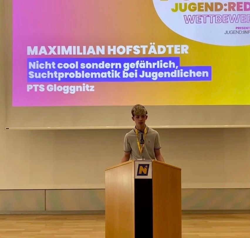 Ein Junge steht an einem Rednerpult mit Mikrofon, hinter ihm steht auf einer Leinwand 'Maximilian Hofstaedter, Nicht cool sondern gefährlich, Suchtproblematik bei Jugendlichen, PTS Gloggnitz.'