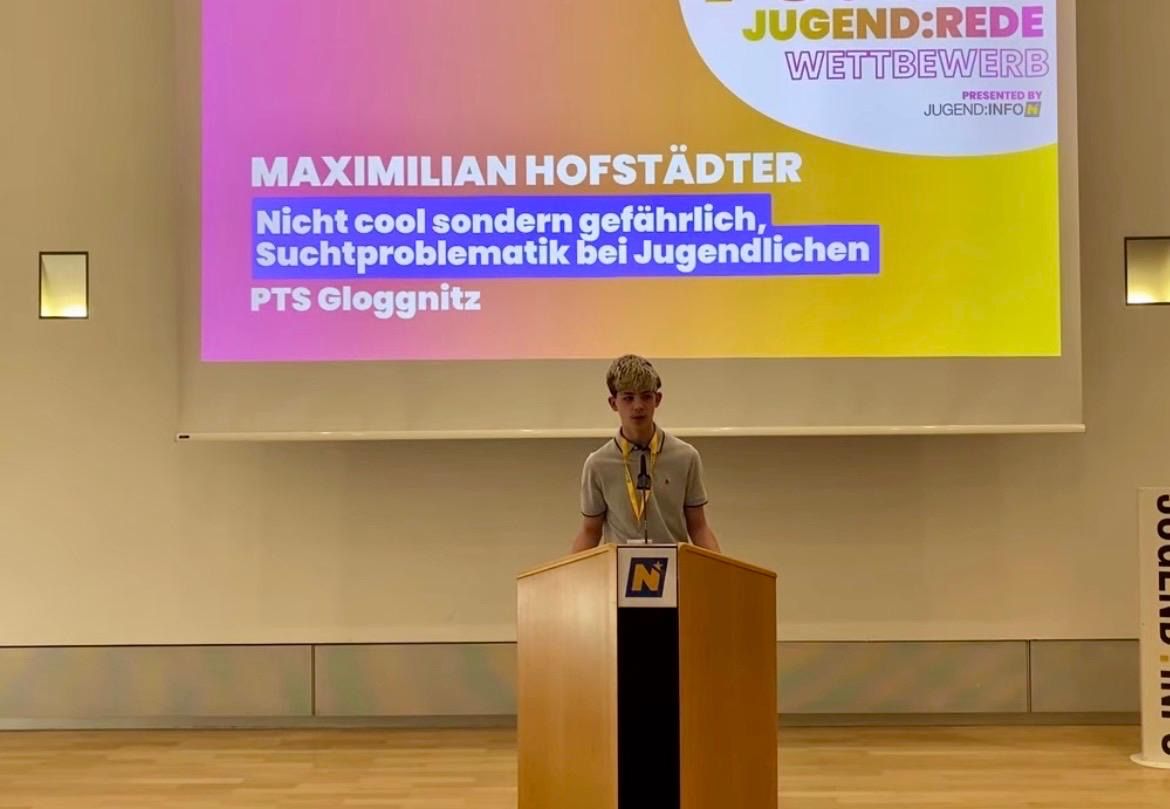 Ein Junge steht an einem Rednerpult mit Mikrofon, hinter ihm steht auf einer Leinwand 'Maximilian Hofstaedter, Nicht cool sondern gefährlich, Suchtproblematik bei Jugendlichen, PTS Gloggnitz.'