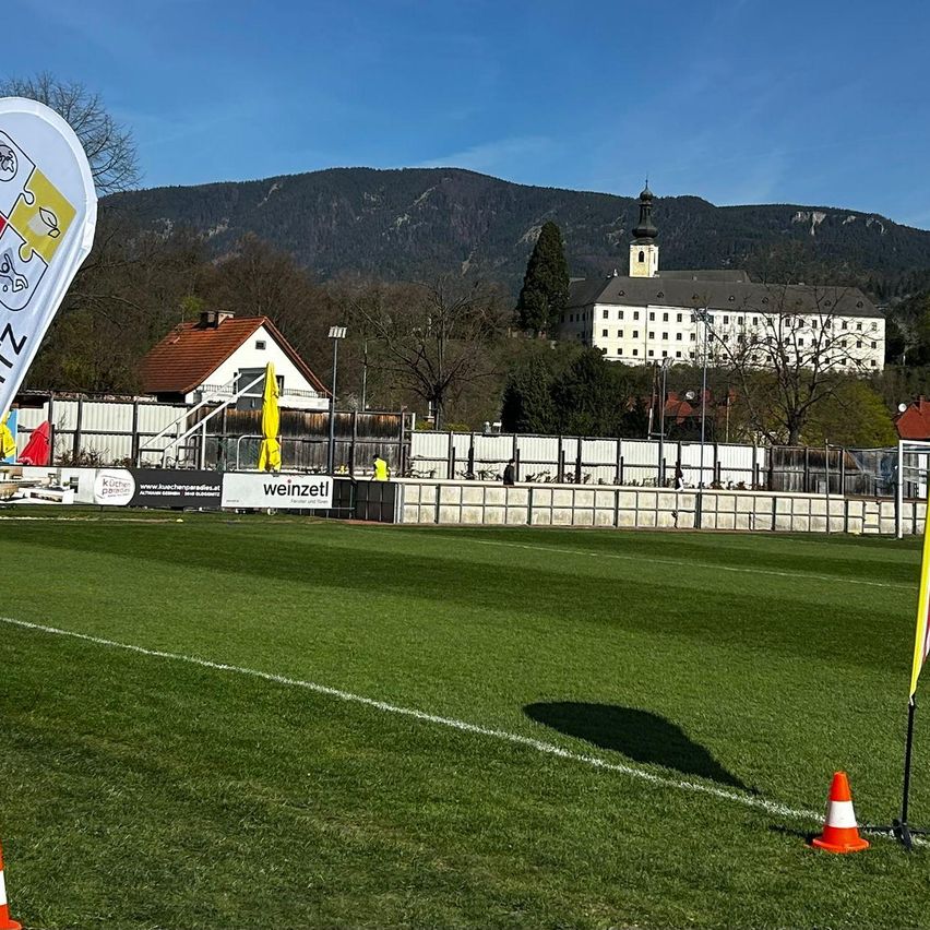 Ein Sportplatz mit einem weißen Banner, Fahnen und Verkehrskegel. Im Hintergrund befindet sich ein großes weißes Gebäude mit Bergen in der Ferne.