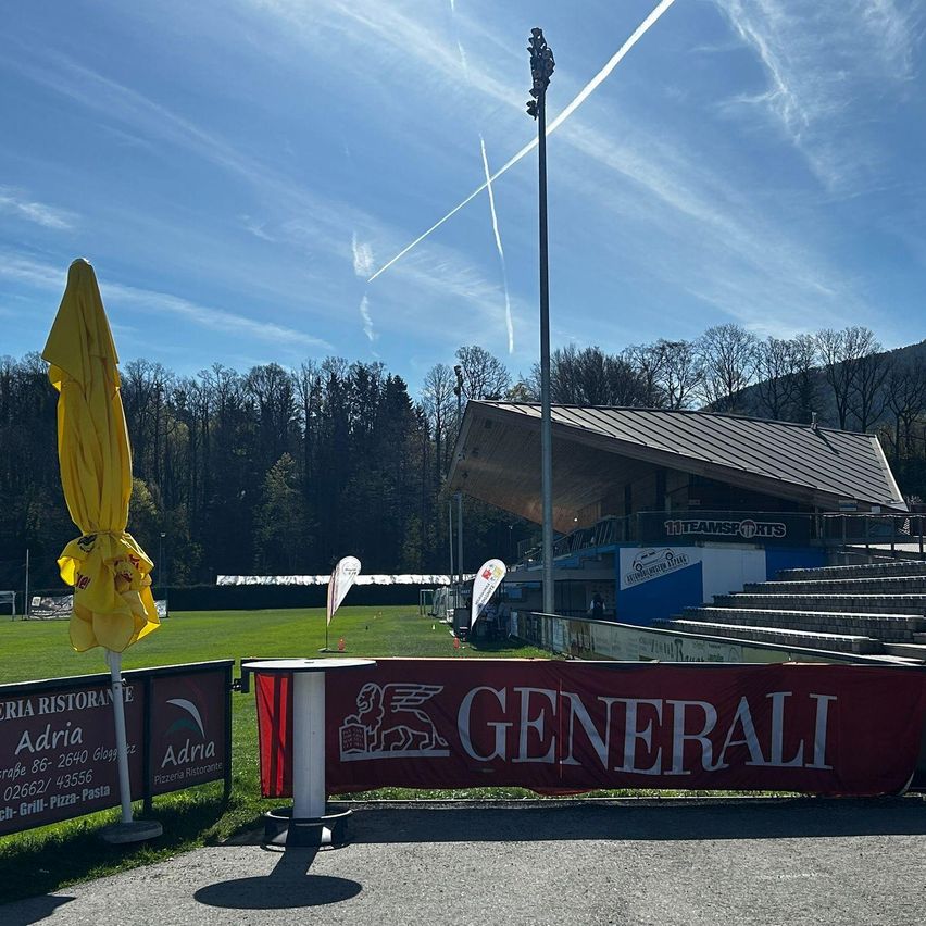 Ein sonniger Tag in einer Sportanlage. Ein gelber Schirm und ein Banner mit der Aufschrift 'Generali' stehen an der Seite. Bäume und Berge sind in der Ferne sichtbar.