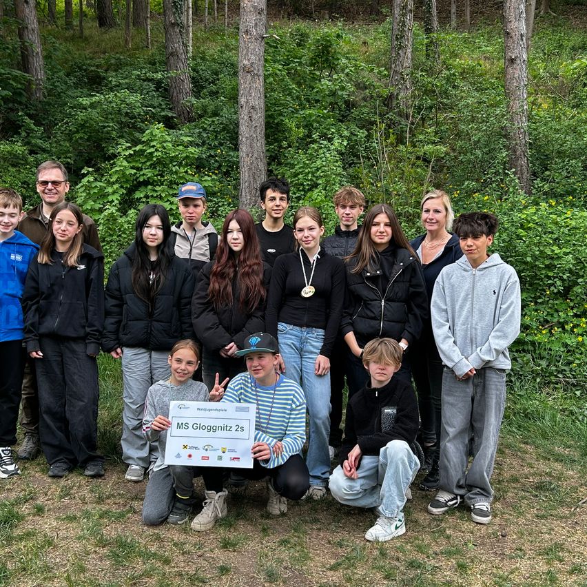 Eine Gruppe von Schülern und Lehrern posiert für ein Foto im Wald. Ein Schüler hält einen großen Scheck für MS Gloggnitz 2s.