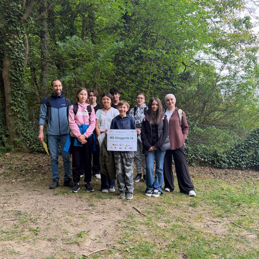 Eine Gruppe von Menschen posiert für ein Foto in einem Wald, wobei sie ein Zertifikat halten. Ein Mann steht links und eine Frau hält ein Banner vor einem Baum.