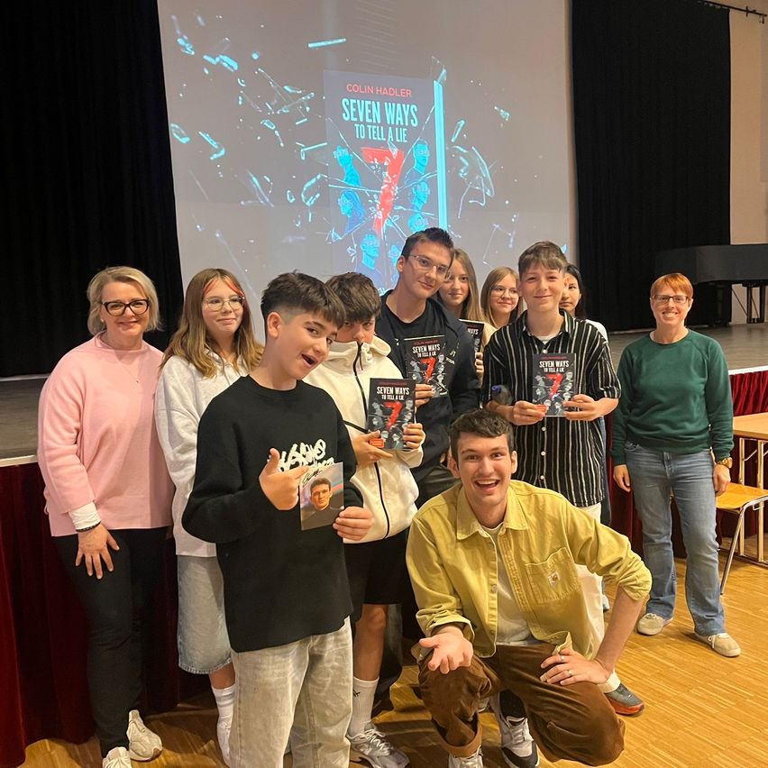 Eine Gruppe von Schülern posiert mit ihren Büchern und einem lächelnden Mann vor einer Leinwand, auf der der Titel 'Sieben Wege, um eine Lüge zu erzählen' zu sehen ist.
