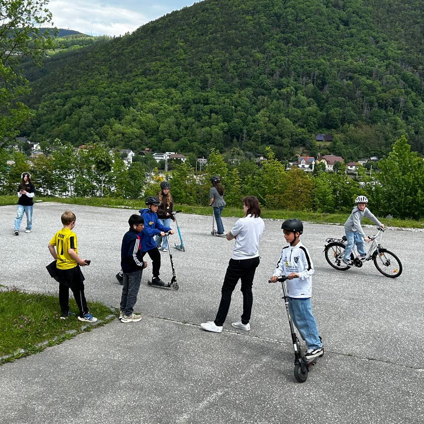 Kinder spielen auf Rollern und Fahrrädern auf einer Straße. Einige tragen Helme. Ein grüner Hügel ist im Hintergrund.