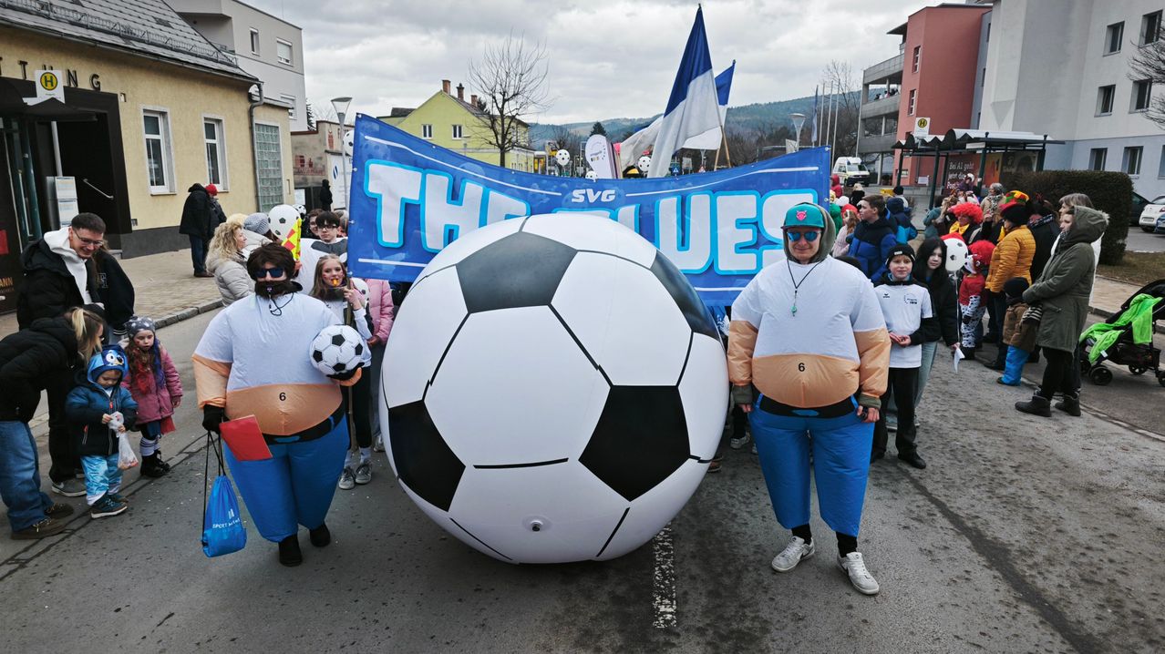 Eine Gruppe von Menschen in Kostümen nimmt mit einem riesigen Fußball an einem Umzug teil, während sie kleinere Fußballbälle halten. Dahinter befindet sich ein Banner in blauen und weißen Farben.