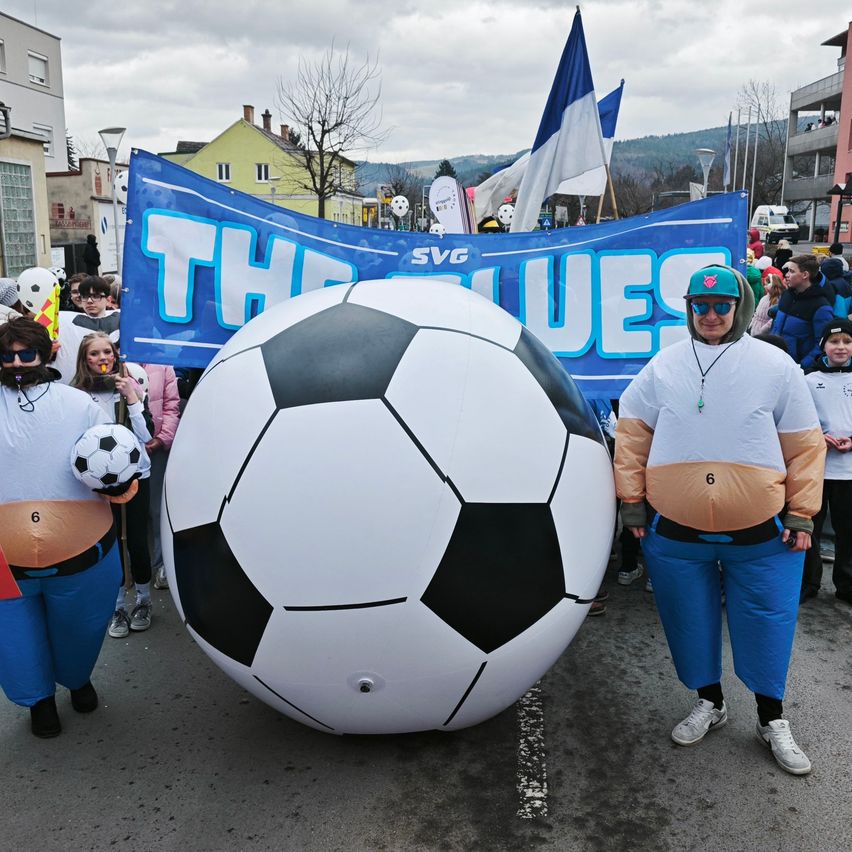 Eine Gruppe von Menschen in Kostümen nimmt mit einem riesigen Fußball an einem Umzug teil, während sie kleinere Fußballbälle halten. Dahinter befindet sich ein Banner in blauen und weißen Farben.