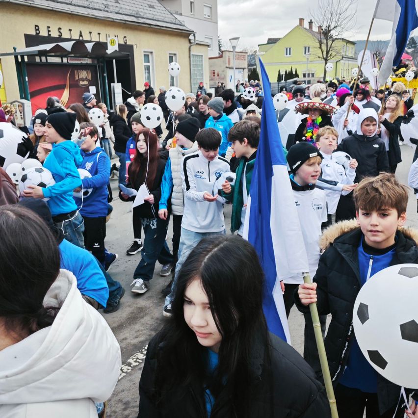 Eine Gruppe von Menschen, überwiegend Kinder, versammelt sich im Freien. Einige halten Fußballbälle, andere tragen Fahnen. Sie sind in Winterkleidung gekleidet und einige tragen Mützen. Ein Gebäude mit dem Schild 'Bestattung' ist im Hintergrund zu sehen.