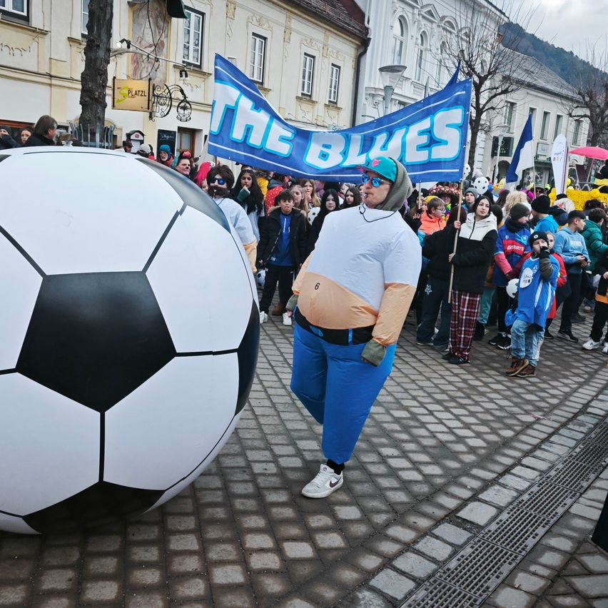 Ein Mann, der als The Blues Maskottchen verkleidet ist, geht an einem riesigen Fußball vorbei, mit einer Menschenmenge hinter ihm auf einer Kopfsteinpflasterstraße.