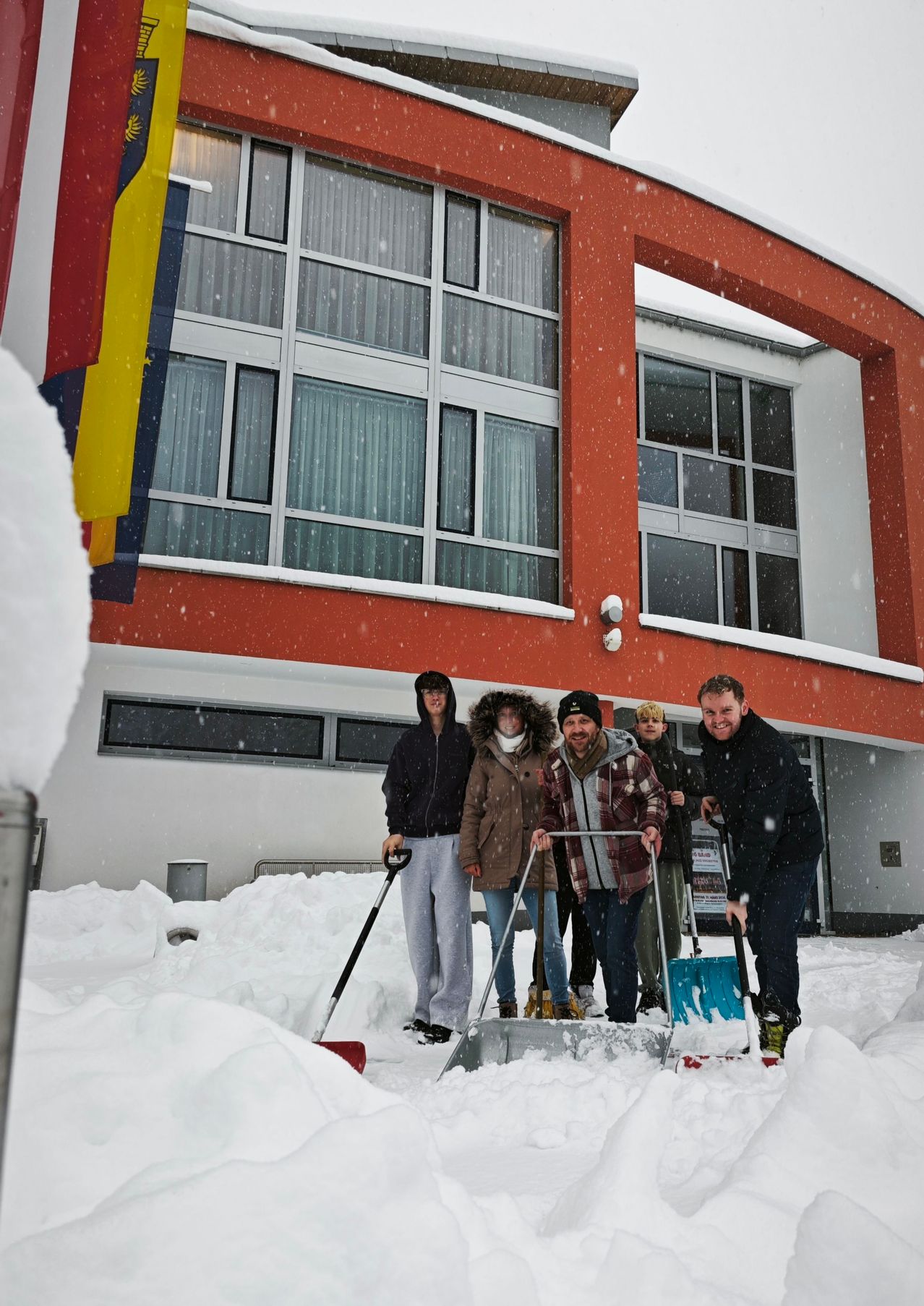 Eine Gruppe von Menschen schaufelt Schnee vor einem roten Gebäude mit vielen Fenstern.