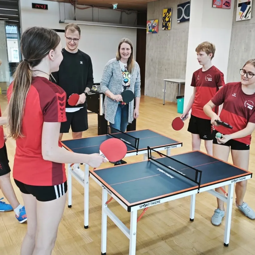 Eine Gruppe von Menschen spielt Tischtennis in einem Raum. Vier von ihnen tragen rote Uniformen und spielen, während die anderen zusehen.