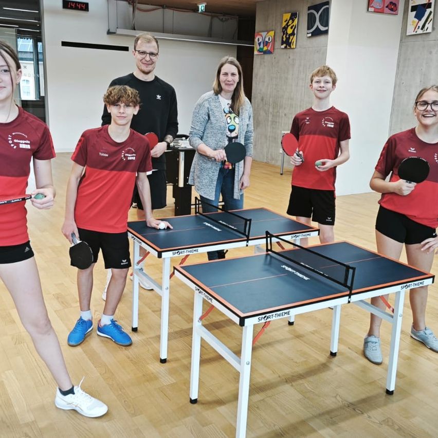 Fünf Personen stehen in einem Raum und posieren möglicherweise für ein Foto. Sie halten alle Tischtennisschläger. Der Raum hat Holzböden und weiße Wände. Die Frau in der Mitte hält einen Schläger in beiden Händen. Es gibt drei Tische mit schwarzen und weißen Oberflächen.