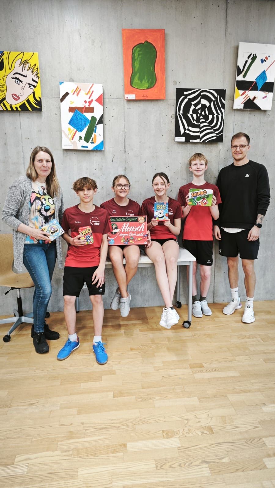 Eine Gruppe von sechs Personen steht und sitzt auf einer Bank und hält Boxen, wahrscheinlich in einem Studio. Hinter ihnen hängen Gemälde an der Wand.