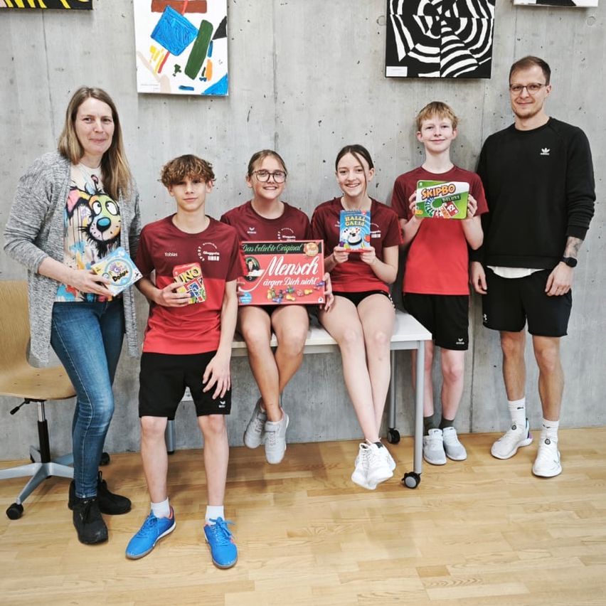 Eine Gruppe von sechs Personen steht und sitzt auf einer Bank und hält Boxen, wahrscheinlich in einem Studio. Hinter ihnen hängen Gemälde an der Wand.