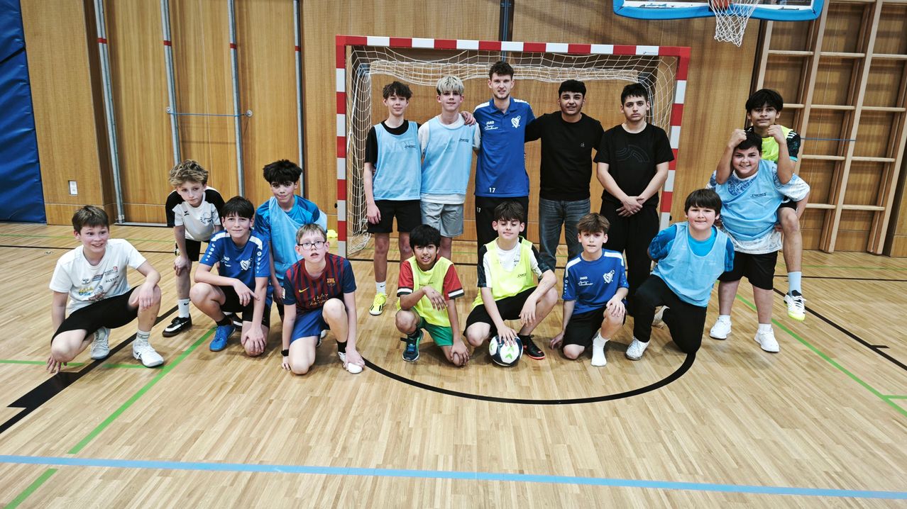 Eine Gruppe von Jungen in Sporttrikots und Shorts posiert für ein Foto in einer Turnhalle, einige knien und einer hält einen Fußball. Ein Tor ist im Hintergrund.