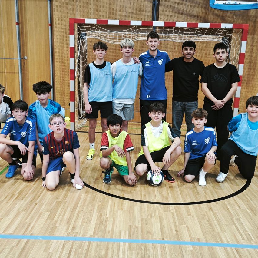 Eine Gruppe von Jungen in Sporttrikots und Shorts posiert für ein Foto in einer Turnhalle, einige knien und einer hält einen Fußball. Ein Tor ist im Hintergrund.