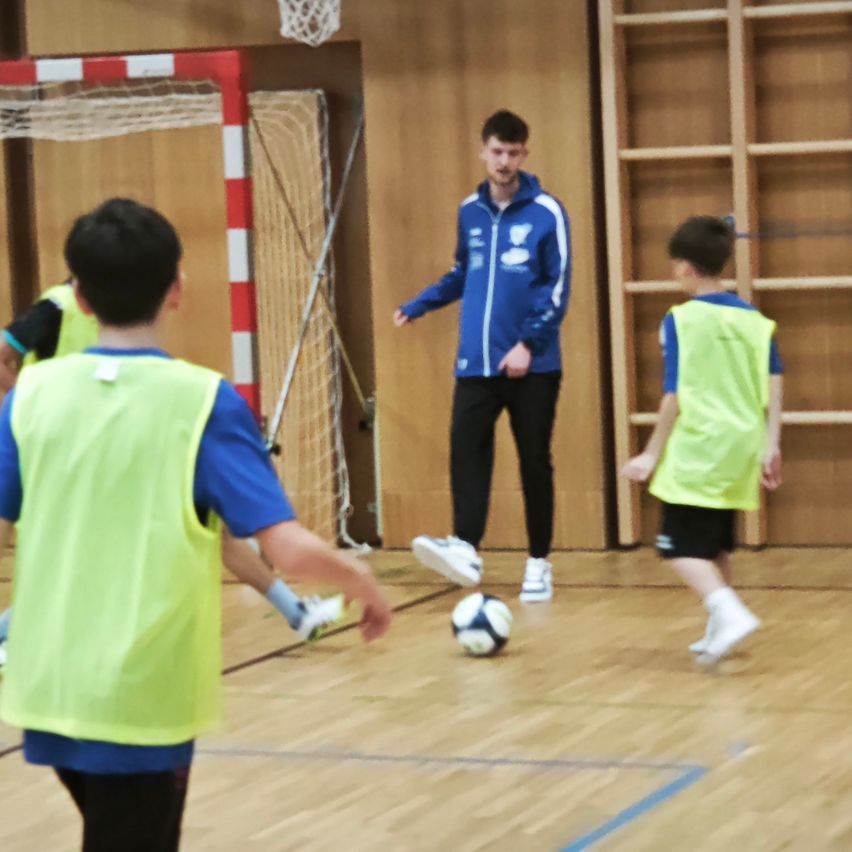Ein Mann in einer blauen Jacke trainiert ein Fußballspiel mit drei Kindern in gelben Westen. Ein Fußball liegt vor ihnen auf dem Boden.